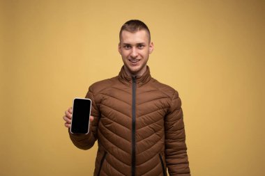 20 'li yaşlarda, sarı arka planda kahverengi ceketli genç bir adam elinde bir cep telefonu tutuyor.
