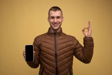 Sarı arka planda kahverengi ceketli genç bir adam, boş ekranlı bir cep telefonu kullanıyor, çalışma alanı gösteriyor ki...
