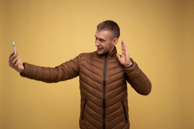 20 'li yaşlarda, kahverengi ceketli genç bir adam, sarı arka planda, cep telefonundan bir video çağrısı alıyor, hoş bir konuşma yapıyor, eliyle selamlıyor.