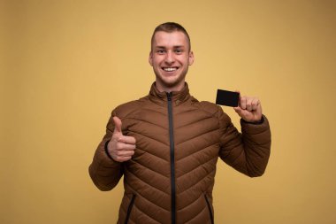Sarı arka planda kahverengi ceketli genç bir adam elinde kredi kartı tutuyordu. Zenginlik Kariyeri Başarı Konsepti