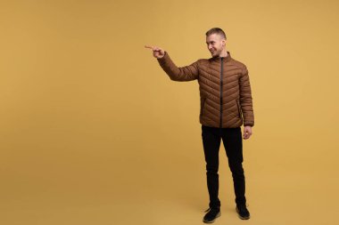 Sarı arka planda, kahverengi ceketli, 20 yaşında, alışılagelmedik çekici genç bir adam, parmağı olan birini işaret ediyor.