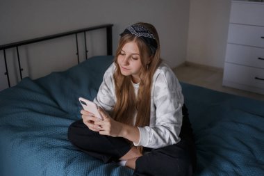 Kafasında bandajla ve uzun sarı saçlarıyla genç bir kız oturma odasında bir yatağa oturuyor, elinde bir telefon tutuyor ve gülüyor.