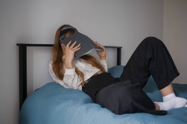 Kafasında bandajla genç 20 'li yaşlarda bir kız ve uzun sarı saçları yüzünü kitapla örten bir yatakta yatıyor.