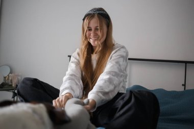 Kafasında bandajla ve uzun sarı saçlarıyla genç bir kız oturma odasında bir yatakta oturmuş bir köpekle oyun oynuyor.