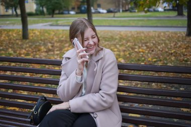 Güzel bir ceketi olan genç bir kız şehirde bir bankta oturuyor, saçları toplanmış, telefonda konuşuyor ve gülüyor.