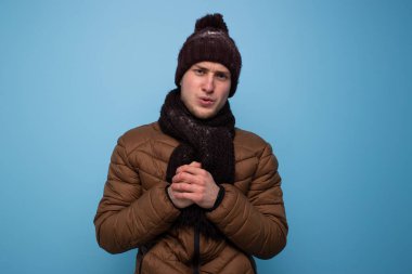 Kahverengi atkı ve kışlık şapkalı genç bir adam mavi arka planda açık kahverengi bir ceket ellerini ısıtıyor.