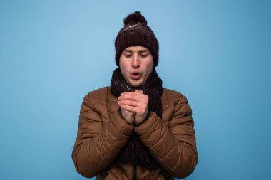 Kahverengi atkı ve kışlık şapkalı genç bir adam mavi arka planda açık kahverengi bir ceket ellerini ısıtıyor.