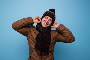 Kahverengi eşarplı, kışlık şapkalı, açık kahverengi ceketli, mavi arka planda kış şapkalı ve gülümseyen genç bir adam.