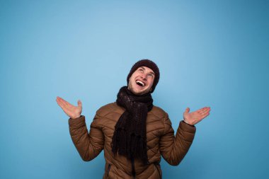 Kahverengi atkı ve kışlık şapkası olan mutlu genç bir adam mavi arka planda açık kahverengi bir ceket gülümsüyor ve avuçlarını üstte gösteriyor.