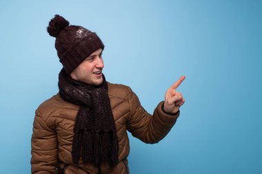 Kahverengi eşarplı ve kışlık şapkalı mutlu genç adam mavi arka planda açık kahverengi bir ceket gülümsüyor ve yan tarafta bir parmağını gösteriyor.