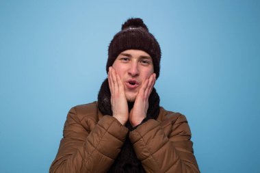 Kahverengi eşarplı ve kışlık şapkalı mutlu genç adam mavi arka planda açık kahverengi bir ceket gülümsüyor ve avuçlarını yanaklarına yaklaştırıyor.