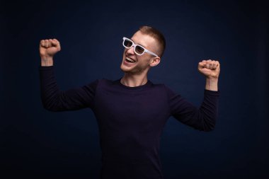 Mavi süveter ve beyaz gözlüklü gülümseyen genç adam mavi bir arka planda poz verir, ve yana bakar ve sevinir