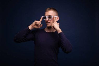 Mavi süveterli ve beyaz gözlüklü gülümseyen genç adam mavi arka planda poz veriyor, gözlüklerini düzeltiyor ve yan tarafa bakıyor.