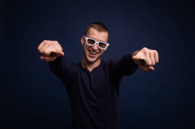 Mavi süveterli ve beyaz gözlüklü gülümseyen genç adam mavi arka planda poz veriyor, parmaklarıyla düz gösteriyor.