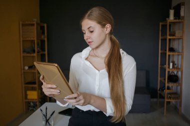 Beyaz gömlekli başarılı bir iş kadını elinde tabletle modern bir ofiste dizüstü bilgisayarı olan beyaz bir masanın yanında duruyor. Kariyer başarısı, başarı ve kişisel büyüme kavramı