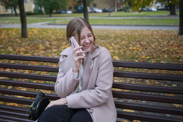Güzel bir ceketi olan genç bir kız şehirde bir bankta oturuyor, saçları toplanmış, telefonda konuşuyor ve gülüyor.