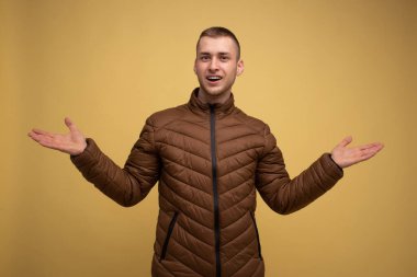 Stüdyo portresi. Sarı arka planda, kahverengi ceketli genç bir adam ellerini sallıyor.