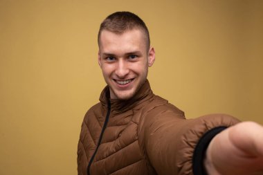 Stüdyo portresi. Sarı arka planda kahverengi ceketli genç bir adam cep telefonundan birinci şahıs selfie çekiyor.