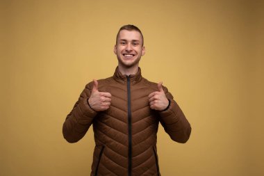 Stüdyo portresi. Sarı arka planda, kahverengi ceketli genç bir adam baş parmağını bir jest olarak gösteriyor.