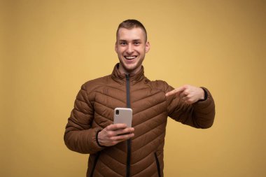 Stüdyo portresi. 20 'li yaşlarda kahverengi ceketli genç bir adam, sarı arka planda, elinde cep telefonu tutuyor, diyor ki, vay canına, zaferler hakkında harika haberler var.