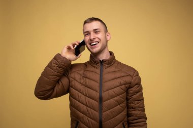 Sarı arka planda kahverengi ceketli genç bir adam, cep telefonundan bir video çağrısı alıyor.