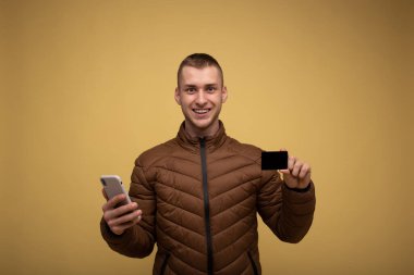 Kahverengi ceketli genç bir adam, sarı arka planda, cep telefonu kullanıyor, elinde bir kredi kartı tutuyor, internetten alışveriş yapıyor.