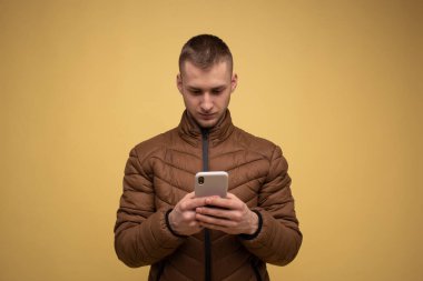 Stüdyo portresi. 20 'li yaşlarda, kahverengi ceketli, sarı arka planda, elinde cep telefonu olan genç bir adam cep telefonu kullanıyor, mesaj yazıyor, sohbet ediyor, SMS gönderiyor.