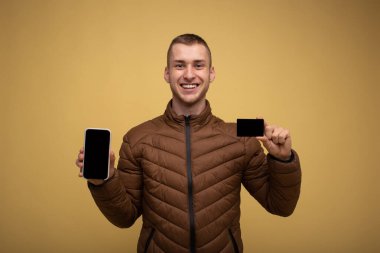 Kahverengi ceketli genç bir adam, sarı arka planda, cep telefonu kullanıyor, elinde bir kredi kartı tutuyor, internetten alışveriş yapıyor.