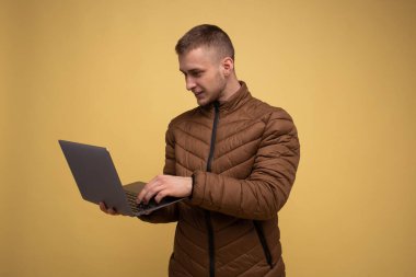 20 'li yaşlarda, kahverengi ceketli genç bir adam, sarı arka planda, dizüstü bilgisayarda iş tutuyor, yazı yazıyor, sohbet ediyor, mesaj yolluyor.