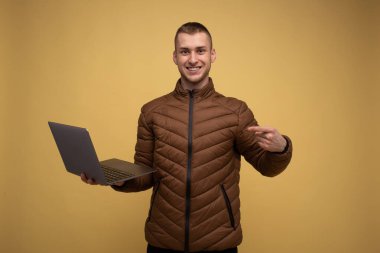 Sarı arka planda kahverengi ceketli genç bir adam dizüstü bilgisayarını tutuyor ve onu işaret ediyor.