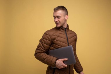 20 'li yaşlarda, kahverengi ceketli genç, sarı arka planda, dizüstü bilgisayarda çalışıyor, dizüstü bilgisayarında çalışıyor, yürüyor, arkasına bakıyor.