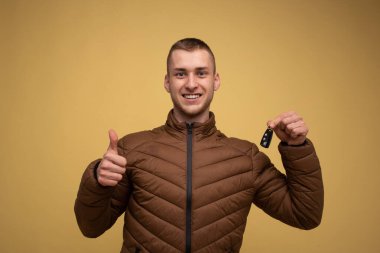 Sarı arka planda, kahverengi ceketli genç bir adam elinde yeni bir araba anahtarı tutuyor.