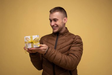 Young 20s man in a brown jacket, on a yellow background, looking at a box with a yellow bow made of a gift ribbon