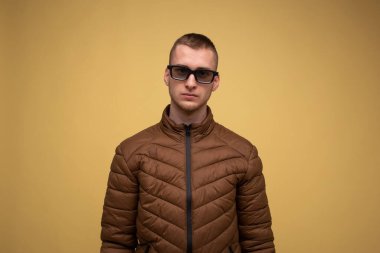 20 'li yaşlarda, kahverengi ceketli, sarı arka planda, üzgün, 3D film izlediği için gözlüklü bir genç.