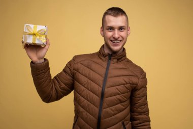 Sarı arka planda kahverengi ceketli genç bir adam, gülümsüyor ve elinde hediye kurdelesinin sarı kurdelesiyle bir kutu tutuyor.