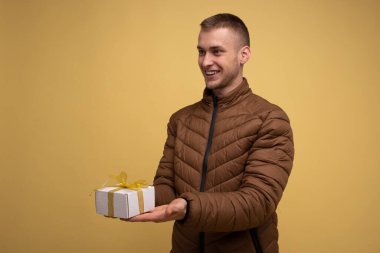 Sarı arka planda kahverengi ceketli genç bir adam, gülümsüyor ve hediye kurdelesinin sarı kurdeleli kutusunu veriyor.