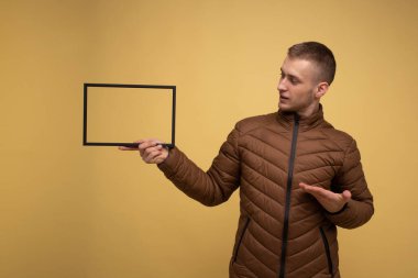 Sarı arka planda, kahverengi ceketli genç bir adam gülümsüyor ve elinde bir fotoğraf çerçevesi tutuyor.