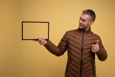 Sarı arka planda, kahverengi ceketli genç bir adam gülümsüyor ve elinde bir fotoğraf çerçevesi tutuyor.