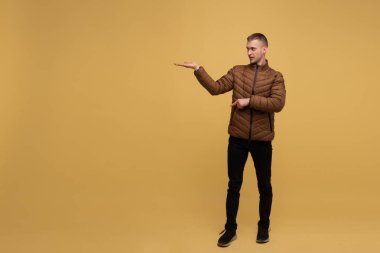 Sarı arka planda, kahverengi ceketli, 20 yaşında, alışılagelmedik çekici genç bir adam, parmağı olan birini işaret ediyor.