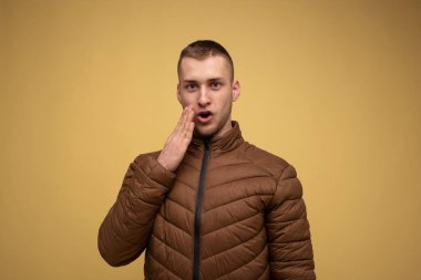 Stüdyo portresi. Sarı arka planda, kahverengi ceketli genç bir adam, yüzünü eliyle kapatıyor, şaşırdığını gösteriyor.