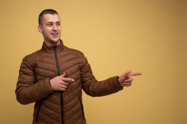 Stüdyo portresi. Sarı arka planda, kahverengi ceketli genç bir adam, parmaklarını bu yöne doğru gösteriyor.