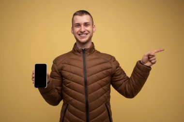 20 'li yaşlarda, kahverengi ceketli, sarı arkaplanlı genç bir adam, elinde bir telefon tutuyor ve parmağını yana çeviriyor.