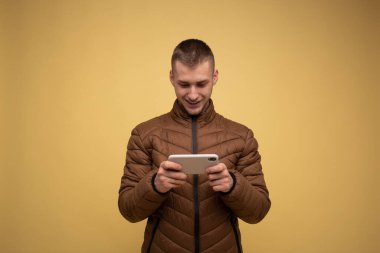 20 'li yaşlarda, kahverengi ceketli genç bir adam, sarı arka planda, elinde bir telefon tutuyor, oyunlar oynuyor, anında habercilerle iletişim kuruyor.