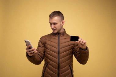 Kahverengi ceketli genç bir adam, sarı arka planda, cep telefonu kullanıyor, elinde bir kredi kartı tutuyor, internetten alışveriş yapıyor.