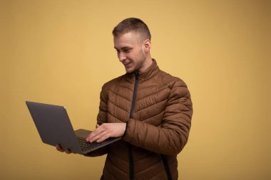 20 'li yaşlarda, kahverengi ceketli genç bir adam, sarı arka planda, dizüstü bilgisayarda iş tutuyor, yazı yazıyor, sohbet ediyor, mesaj yolluyor.