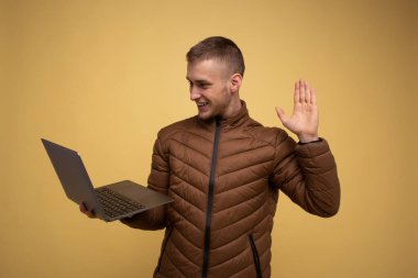 20 'li yaşlarda, kahverengi ceketli, sarı arka planda bir dizüstü bilgisayar tutan, iş, video konferans, el ele selamlaşmalar yapan genç bir adam.