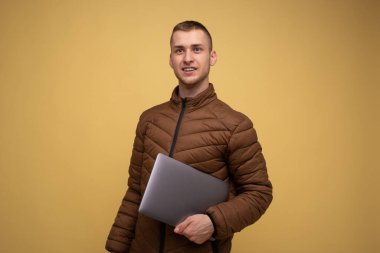 20 'li yaşlarda, kahverengi ceketli genç, sarı arka planda, dizüstü bilgisayarda çalışıyor, laptopta çalışıyor, yürüyor, ileriye bakıyor.