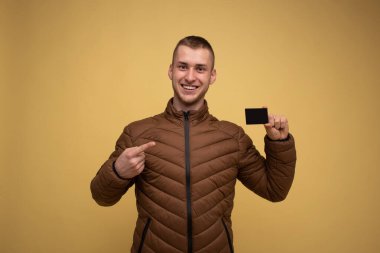 20 'li yaşlarda, kahverengi ceketli, sarı arka planda elinde bir kredi kartı tutan, parmağını gösteren genç bir adam. Zenginlik Kariyeri Başarı Konsepti