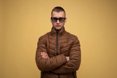 20 'li yaşlarda, kahverengi ceketli, sarı arka planda, üzgün, 3D film izlediği için gözlüklü bir genç.