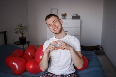 Beyaz tişörtlü bir adam kalp şeklinde balonlar arasında bir kalp gösteriyor, Sevgililer Günü için.